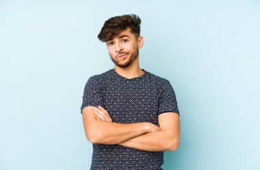 Mavi arka planda izole edilmiş, hoşnutsuzca kaşlarını çatmış, kollarını kavuşturmuş genç bir Arap..