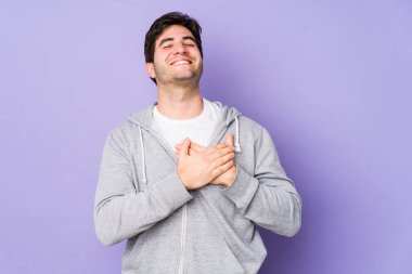 Mor arka planda izole olmuş genç adam gülüyor, ellerini kalbe koyuyor, mutluluk kavramını koruyor..