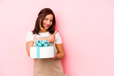 Orta yaşlı Latin bir kadın pembe arka planda izole edilmiş bir pasta tutuyor gülümsüyor ve boş alanda bir şeyler gösteriyor..