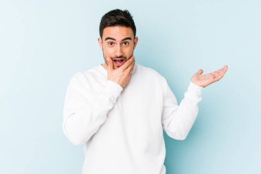 Mavi arka planda izole edilmiş genç beyaz bir adam avucunun içinde fotokopi alanı tutuyor, el üstünde tutuyor. Şaşırmış ve mutlu..