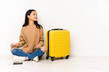 Genç Çinli gezgin kadın, bavuluyla yerde oturuyor. Sola bakıyor, yandan bakıyor..