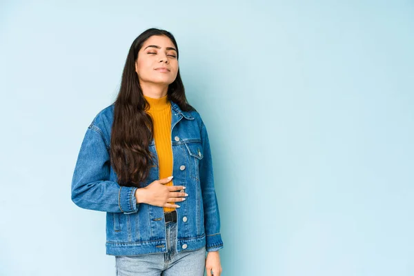 Young indian woman isolated on blue background touches tummy, smiles gently, eating and satisfaction concept.