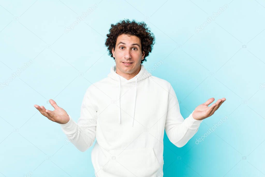 Young sporty man against a blue background doubting and shrugging shoulders in questioning gesture.