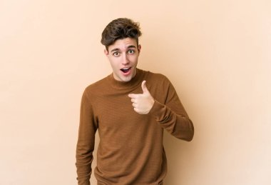 Beyaz tenli genç adam bej arka planda izole edildi. Parmakla göstererek şaşırdı, gülümsedi..