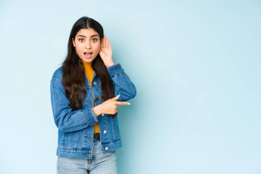 Genç Hintli bir kadın mavi arka planda bir dedikoduyu dinlemeye çalışıyor..
