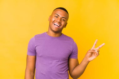 Sarı arka planda izole edilmiş genç Latin adam neşeli ve kaygısız parmakları olan bir barış sembolü gösteriyor..