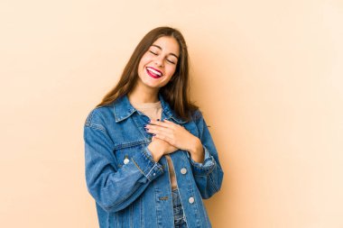 Beyaz tenli genç bir kadın bej arka planda izole edilmiş. Gülüyor, elini kalbe koyuyor, mutluluk kavramını koruyor..