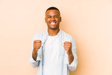 Genç latin adam bej arka planda izole edilmiş tasasız ve heyecanlı tezahüratlar yapıyor. Zafer konsepti.