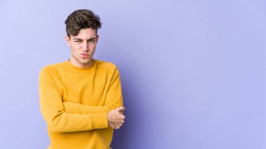 Mor arka planda izole edilmiş genç beyaz adam hoşnutsuzca kaşlarını çatmış, kollarını kavuşturmuş..