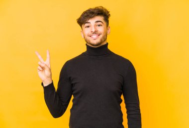Sarı arka planda izole edilmiş genç bir Arap. Neşeli ve tasasız. Parmakları olan bir barış sembolü gösteriyor..