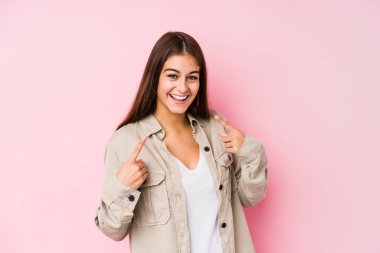 Pembe arka planda poz veren beyaz bir kadın gülümsüyor, parmaklarını ağzına doğrultuyor..