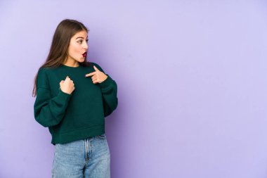 Mor arka planda izole edilmiş beyaz bir kadın. Parmakla göstererek şaşırmış, geniş bir gülümsemeyle..