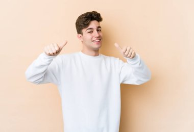 Beyaz tenli genç adam bej arka planda izole edilmiş. Baş parmağını kaldırıyor, gülümsüyor ve kendinden emin..