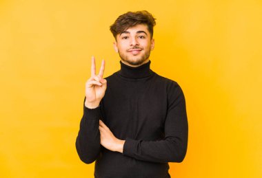 Sarı arka planda izole edilmiş genç bir Arap adam iki numarayı parmaklarıyla gösteriyor..