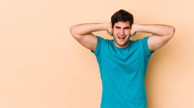 Genç adam bej arka planda izole edilmiş kulakları elleriyle kapatıyor çok fazla ses duymamaya çalışıyor..