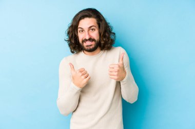 Genç, uzun saçlı adam mavi bir arka planda izole edilmiş. Baş parmağını kaldırıyor, gülümsüyor ve kendinden emin..