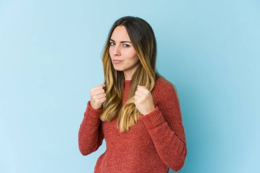 Mavi arka planda izole edilmiş genç beyaz kadın yumruğunu kameraya gösteriyor, agresif yüz ifadesi..