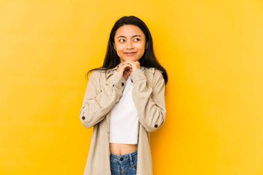 Sarı arka planda izole edilmiş genç Çinli kadın ellerini çenesinin altında tutuyor ve mutlu bir şekilde yan tarafa bakıyor..