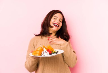 Orta yaşlı Latin bir kadın waffle tutuyor yalnız kahkahalar atıyor elini göğsüne koyuyor.