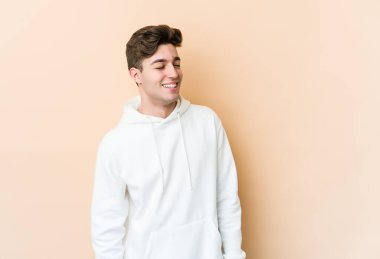 Beyaz tenli genç adam bej arka planda izole edilmiş rahat ve mutlu bir şekilde gülüyor, boynunda dişlerini gösteriyor..