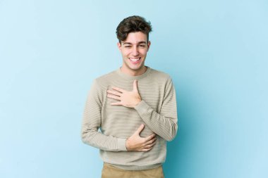 Mavi arka planda izole edilmiş genç beyaz adam mutlu bir şekilde gülüyor ve ellerini karnının üstünde tutarak eğleniyor..