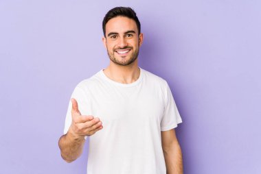 Mor arka planda izole edilmiş genç beyaz adam selamlama jesti olarak kameraya elini uzatıyor..