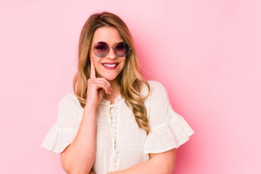 Pembe arka planda gözlüklü genç beyaz kadın gülümsüyor ve kendinden emin, elleriyle çenesine dokunuyor..