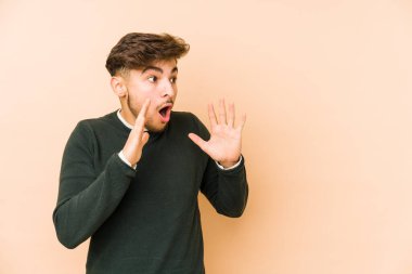 Bej bir arka planda izole edilmiş genç bir Arap yüksek sesle bağırıyor, gözleri açık ve elleri gergin tutuyor.