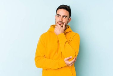 Mavi arka planda izole edilmiş genç beyaz adam şüpheli ve şüpheci bir ifadeyle yana bakıyor..