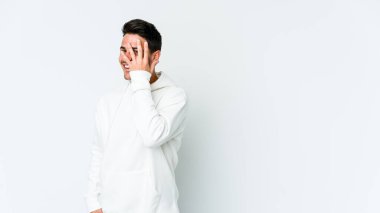 Beyaz arka planda izole edilmiş genç beyaz bir adam kameraya parmaklarıyla göz kırpıyor, yüzünü kapatmaktan utanıyor..