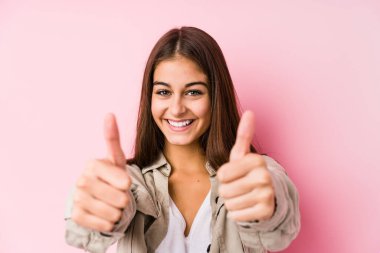 Pembe arka planda poz veren beyaz bir kadın. Başparmakları havada. Bir şey için tezahürat yapıyor..