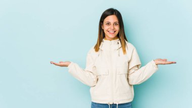 Mavi arka planda soyutlanmış genç bir kadın kollarıyla ölçek oluşturuyor, mutlu ve kendinden emin hissediyor..