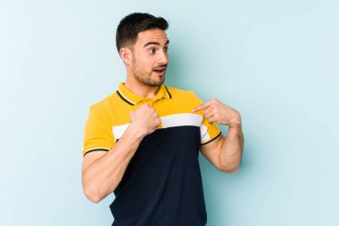 Mavi arka planda izole edilmiş genç beyaz adam parmağını göstererek şaşırdı, geniş bir gülümsemeyle..