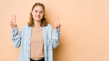 Bej arka planda izole edilmiş genç beyaz kadın zafer işareti gösteriyor ve gülümsüyor..