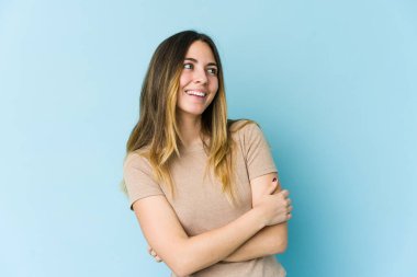 Mavi arka planda izole edilmiş genç beyaz kadın kollarını kavuşturarak gülümsüyor..