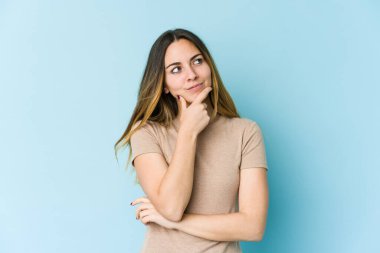 Young caucasian woman isolated on blue background thinking and looking up, being reflective, contemplating, having a fantasy. clipart