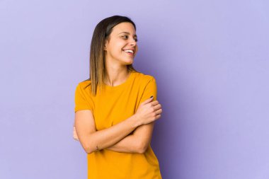 Mor arka planda soyutlanmış genç bir kadın kollarını kavuşturarak gülümsüyor..