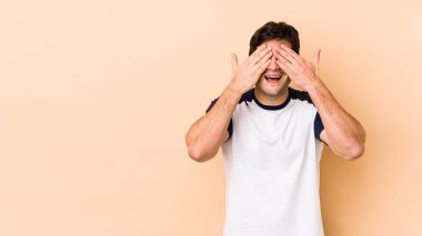 Bej arka planda izole edilmiş genç adam elleriyle gözlerini kapatıyor, geniş bir gülümsemeyle bir sürpriz bekliyor..