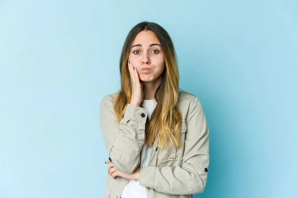Young caucasian woman isolated on blue background blows cheeks, has tired expression. Facial expression concept.