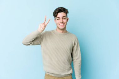 Kafkasyalı genç adam mavi arka planda izole edilmiş neşeli ve kaygısız parmaklarıyla barış sembolü gösteriyor..