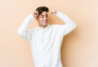 Beyazlı genç adam bej arka planda izole edilmiş özel bir günü kutluyor, atlıyor ve enerjiyle kollarını kaldırıyor..