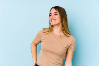 Genç beyaz kadın mavi arka planda izole edilmiş rahat ve mutlu bir şekilde gülüyor, boynunda dişlerini gösteriyor..