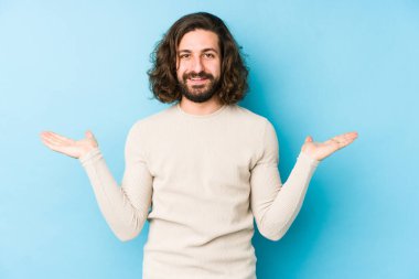 Mavi arka planda izole edilmiş genç, uzun saçlı adam kollarıyla ölçeği yapıyor, mutlu ve kendinden emin hissediyor..