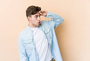 Beyazlı genç bir adam bej arka planda izole edilmiş, uzaklara bakıyor ve elini alnına koyuyor..