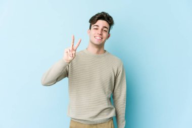 Mavi arka planda izole edilmiş genç beyaz adam zafer işareti gösteriyor ve gülümsüyor..