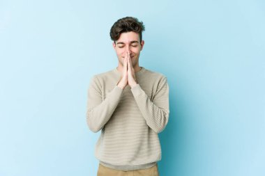Mavi arka planda izole edilmiş genç beyaz bir adam el ele tutuşup, ağzının kenarında dua ediyor, kendinden emin..