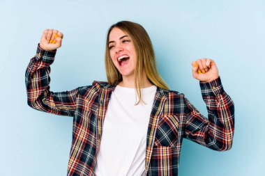 Mavi arka planda izole edilmiş genç beyaz kadın özel bir günü kutluyor, atlıyor ve enerjiyle kollarını kaldırıyor..