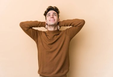 Beyazlı genç bir adam bej arka planda izole edilmiş. Kendine güveniyor, elleri başının arkasında..