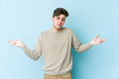 Kafkasyalı genç adam mavi arka planda izole edilmiş sorgulama jestinde şüphe ediyor ve omuz silkiyor..
