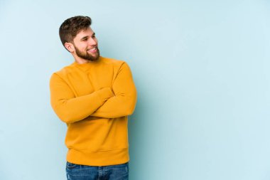 Mavi arka planda izole edilmiş genç beyaz adam kollarını kavuşturarak gülümsüyor..
